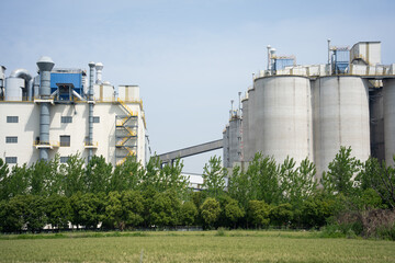 Fototapeta premium Modern industrial factory storage tanks hidden behind green trees under blue sky