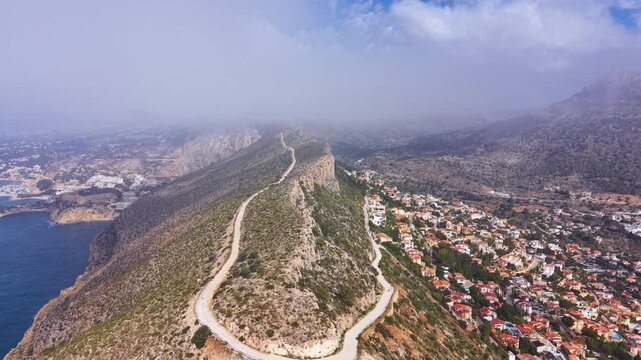 Aerial view of Morro de Toix mountain ridge in Calpe, Spain, with winding dirt road, Mediterranean coastline, misty clouds and residential town below