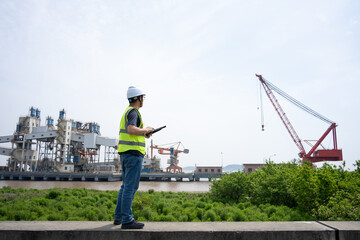 Fototapeta premium Engineer standing at port terminal holding tablet computer with cranes and industrial machinery in background
