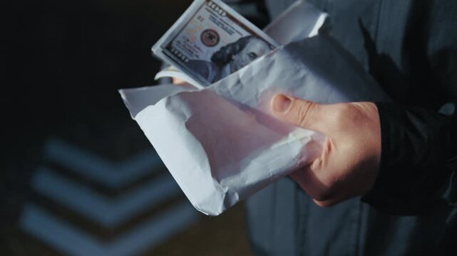 Close-up of male hands counting a stack of hundred-dollar bills from a white envelope at night