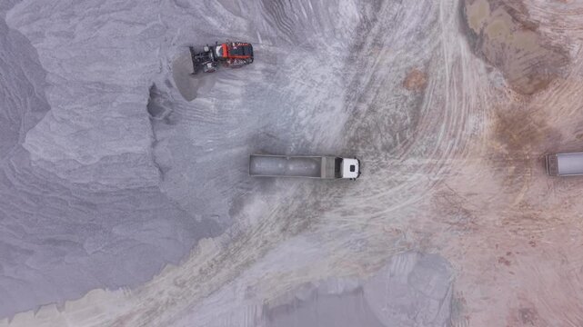 Overhead view of a dump truck being loaded by an orange excavator in a large open pit quarry with white mineral deposits and dirt roads