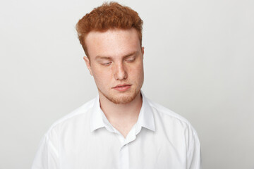 Obraz premium Young man with red hair and freckles wearing white shirt looks down with closed eyes on plain light background. Concept of contemplation, calmness, and introspection in studio portrait.