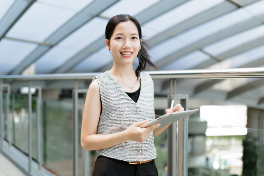 A smiling woman stands indoors holding a tablet and stylus, with a modern glass ceiling and railing in the background.