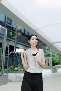 A cheerful woman stands outdoors in a modern urban setting, holding a tablet and smiling confidently.