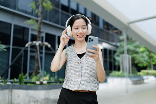 A young woman wearing headphones smiles while using her smartphone outdoors near a modern building.