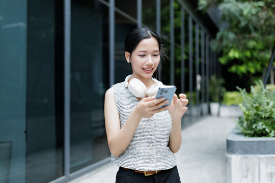 A young woman outdoors smiles while using her smartphone, wearing headphones around her neck near a modern building.