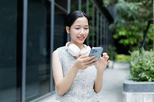 A young woman outdoors smiles while using her smartphone, with headphones resting around her neck near modern glass buildings.