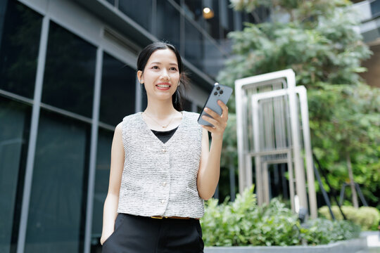 A smiling woman stands outdoors in an urban setting, holding a smartphone and looking away, surrounded by modern architecture and greenery.