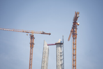 Fototapeta premium Two concrete bridge towers and yellow cranes against a clear blue sky background