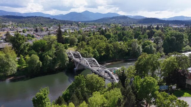 Grants Pass Oregon with white concrete arch bridge crossing Rogue River, lush green tree canopy, charming small town sprawl and forested blue mountain ranges under sunny cloudy sky. Aerial wide shot.