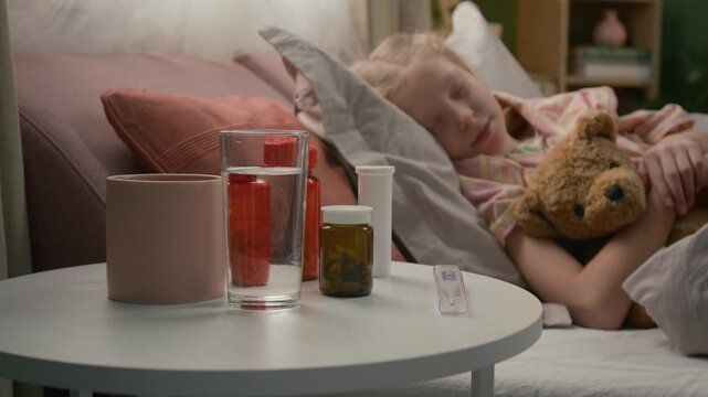 Selective focus shot of mug, glass of water, thermometer and bottles with pills on side table in foreground, sick kid sleeping in bed in background