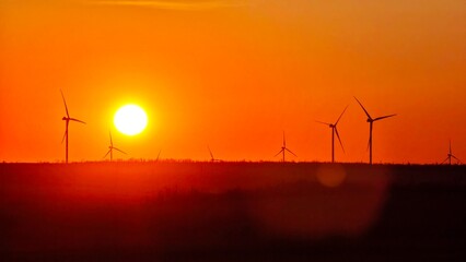 Fototapeta premium A vibrant sunset over a landscape with several wind turbines silhouetted against the orange sky. The scene captures renewable energy and natural beauty.