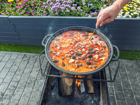 Traditional Thai Tom Yum soup cooking in a large kazan over an open fire outdoors, hand stirring spicy seafood soup with mushrooms in a garden setting