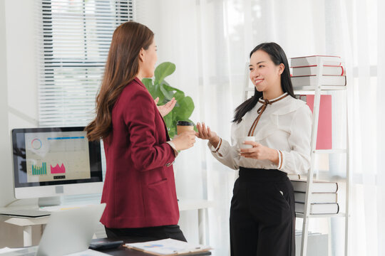 Office Collaboration: Two professional women engage in a friendly discussion while holding coffee cups in a modern office setting, with a computer displaying charts and a bookshelf in the background.