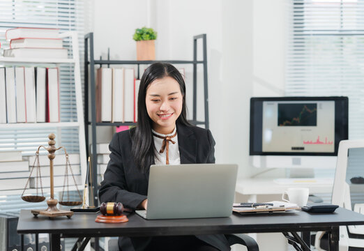 Lawyer Working in Office: A professional female lawyer in a black suit sits at her desk in a modern office, typing on a laptop, with legal scales and a gavel nearby.