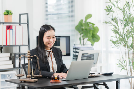 Asian Lawyer at Work: An Asian female lawyer, dressed in formal attire, smiles while working on her laptop at her office desk, surrounded by legal paraphernalia such as scales of justice and a gavel.