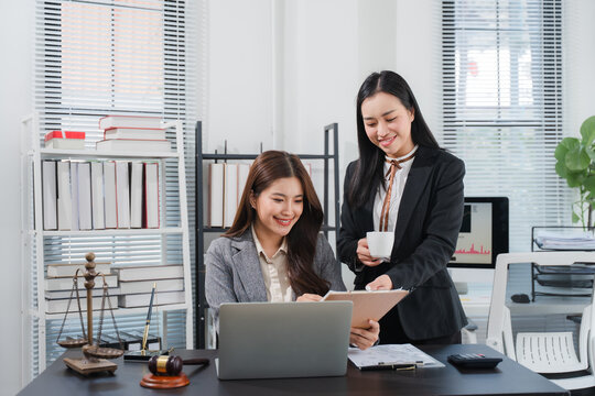 Legal Partnership: Two professional women, lawyers or paralegals, collaborate over documents and a laptop in a well-lit office setting. One holds a cup of coffee.
