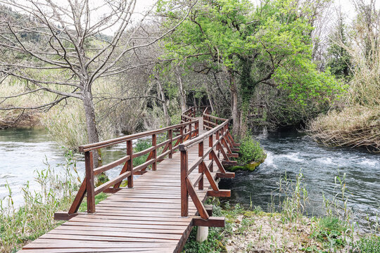 Curved wooden bridge above river in forest landscape. Scenic walkway in natural park. Peaceful outdoor travel destination with flowing water and green trees. Hiking trail in Croatia nature