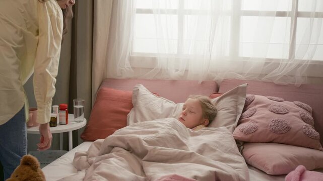 Caring mother bringing hot tea for sick sleeping gen alpha child, adjusting her blanket and pillow, kissing daughter on forehead