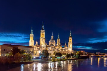 Fototapeta premium Captivating evening panorama of Zaragoza, Spain, featuring the illuminated Basilica del Pilar and the Ebro River. Vibrant city lights reflecting in the water during the blue hour