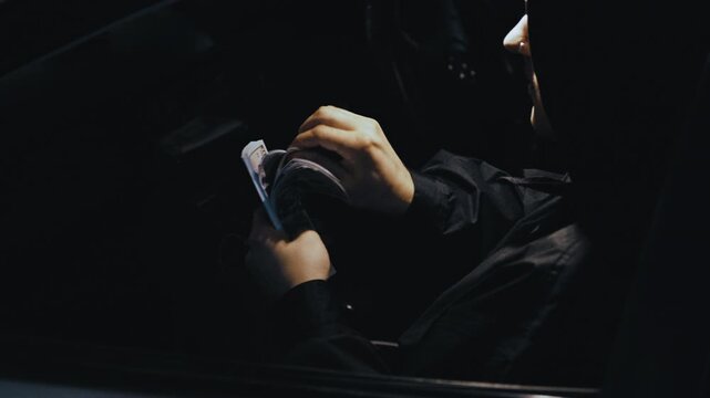 A shady criminal in a hood counts a large amount of US currency inside a car at night. 