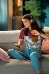 Fototapeta premium A woman sits on a couch, wearing headphones and smiling. She checks her phone and moves to the beat of the music. The room is filled with soft light and decor.