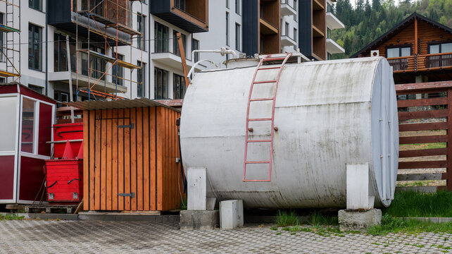 Large industrial storage tank with ladder stands near construction site and wooden cabin