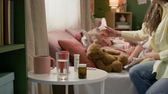 Selective focus view of woman putting glass of water and pill bottle on white bedside table, patting sick kid on head and kissing her