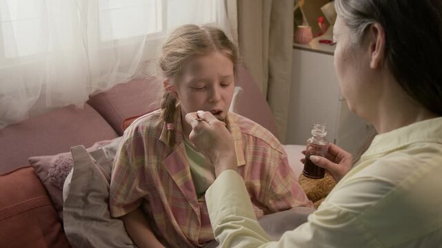 Over the shoulder shot of mother pouring liquid medicine onto spoon and putting it into mouth of sick gen alpha child suffering from cough