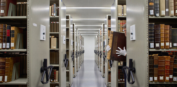 Archivist in protective gloves retrieving historical book from dense mobile shelving units in library storage with archivist literature research education collection organization university
