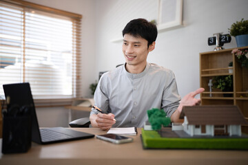 Fototapeta premium Asian Male Architect Presenting House Model During Video Call at Modern Office Desk via laptop, real estate and design concepts