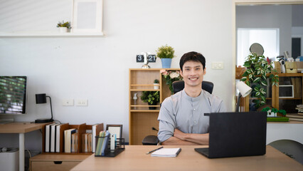 Fototapeta premium Young Asian Businessman Sitting at Desk with arms crossed and Laptop in Modern Home Office with Confident Expression, remote work and business concepts