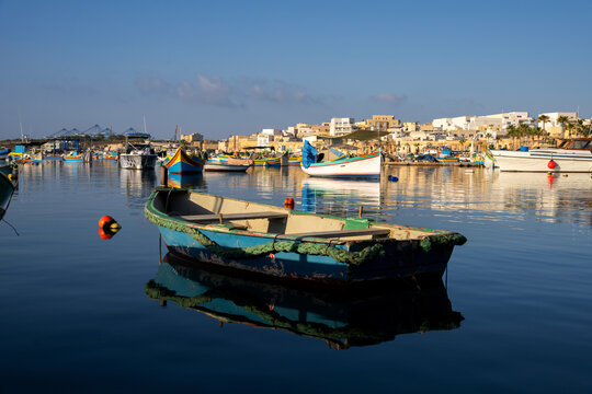Traditional wooden fishing boat with reflection in calm water of Marsaxlokk harbor, Malta, with colorful luzzu boats and village waterfront in background at sunny morning.