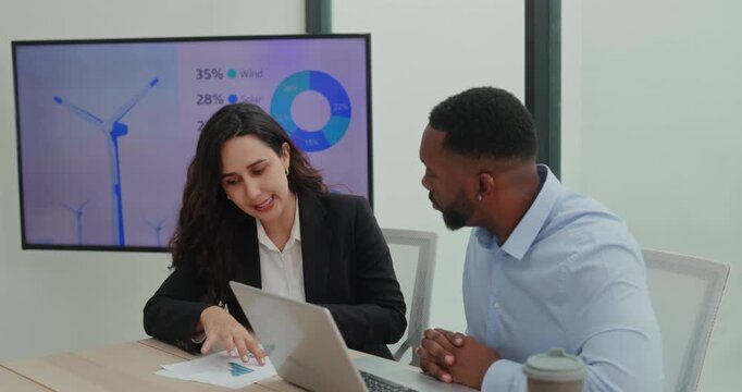 Portrait of Hispanic lady discusses renewable energy report with African American colleague in meeting room. Analyst gestures over documents and explains chart