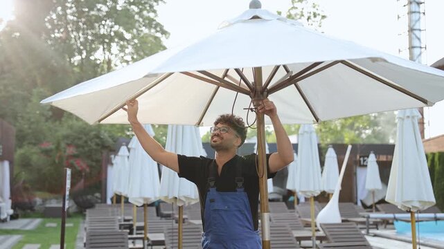 Resort employee wearing overalls and glasses is seen unfolding a large sun umbrella in anticipation of a bustling day ahead at the pool area