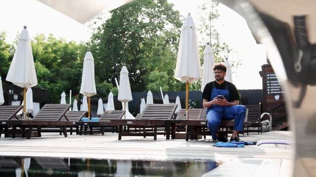 Young man working as a pool technician taking a break, sitting and using smartphone at a sunny outdoor swimming pool