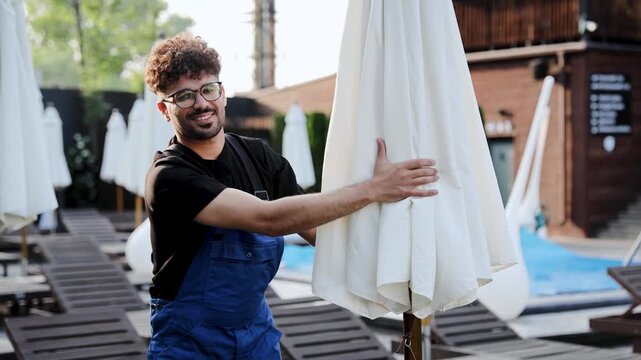 Resort maintenance worker preparing the pool area for guests, opening sunshade umbrellas and arranging sun loungers for the summer