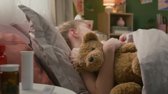 Rack focus view of bottles with pills on side table, and sick girl coughing and turning over on her left side while sleeping in bed with plush bear