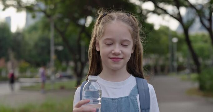 Portrait of pretty junior schoolgirl holds water bottle in green city park. Little girl elementary student with drink stands on town street. Healthy children