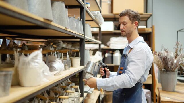 Male small business owner in a denim apron using a handheld barcode scanner to check inventory of metal planters on shelves in a retail home decor and garden boutique shop. Managing store stock.