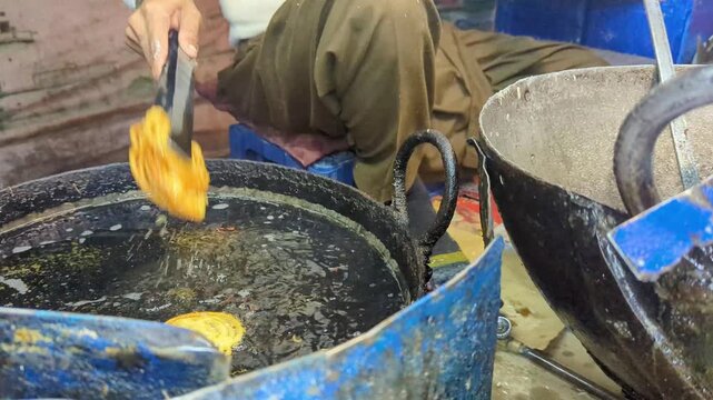 Street vendor using a metal spatula to lift crispy golden jalebi sweets from a large black deep frying pan filled with hot oil at a market