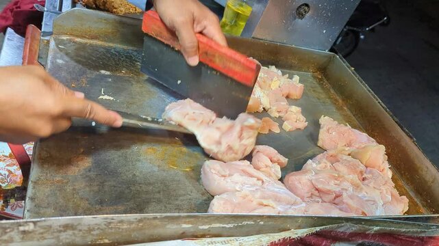 Street vendor using a sharp knife to chop raw chicken breast meat into small pieces on a hot metal griddle for cooking delicious fast food meals