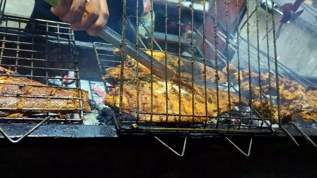 Street chef using metal tongs to flip fresh fish seasoned with spices on a hot charcoal grill at night during a lively traditional outdoor food market