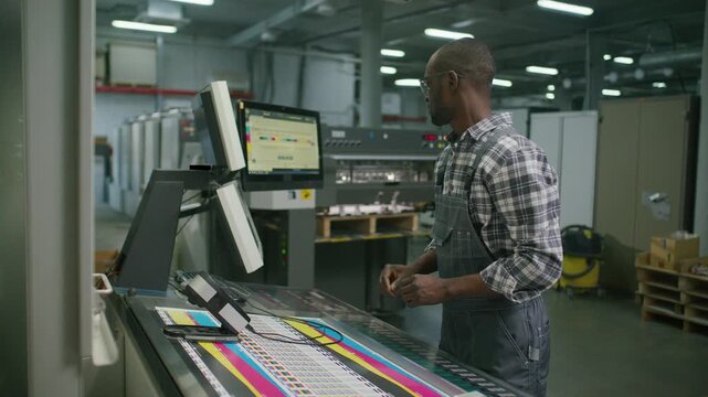 Black male worker in overalls measuring color accuracy with spectrophotometer tool, then using digital monitor screen to operate machine in modern printing house