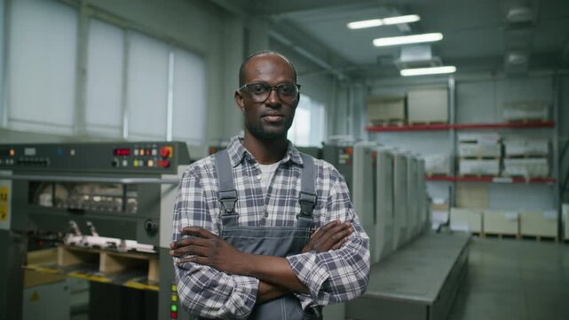 African American male technician in overalls and glasses posing confidently next to automated offset printing press inside printing house. Video portrait