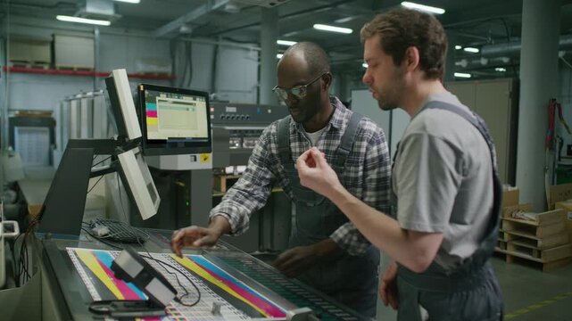 Two multiethnic male workers checking color quality with spectrophotometer and adjusting settings on industrial press machine at modern printing factory