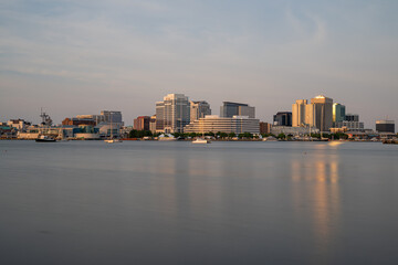 Fototapeta premium Norfolk, Virginia, skyline reflecting on the calm waters of the Elizabeth River during a golden hour sunset, showing high-rise buildings and boats moored along the downtown waterfront.