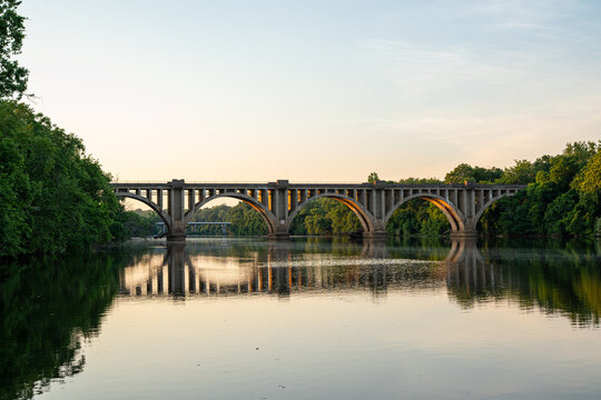 Fredericksburg, Virginia, features the historic concrete arch railroad bridge spanning across the calm, reflective waters of the Rappahannock River surrounded by dense green trees at sunset.