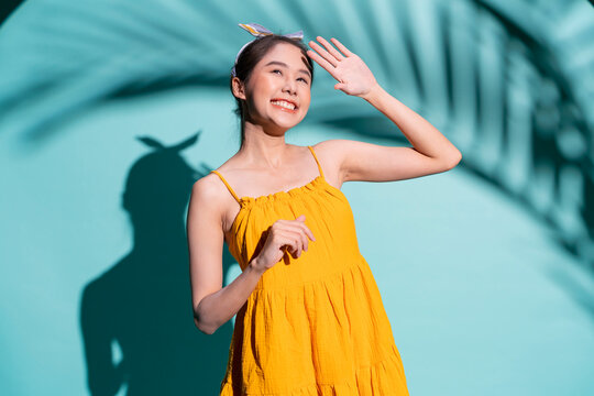 beautiful cheerful young Asian woman in a vibrant yellow swimsuit and sunglasses poses playfully, raising her hands to block the bright summer sunshine while creating a dramatic art shadow