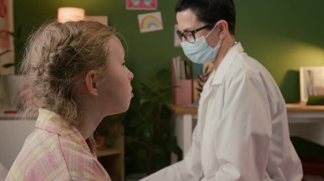 Rack focus shot of short-haired mature female doctor with face mask palpating neck of gen alpha child and examining her throat using spatula